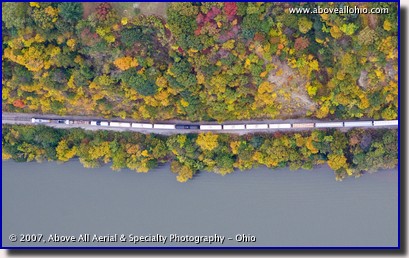 A vertical aerial image of a freight train between a river and fall colored trees in PA